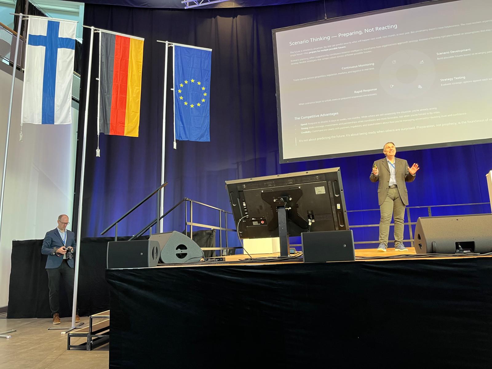 A speaker on stage beside Finnish, German, and European Union flags, presenting a slide about scenario thinking.