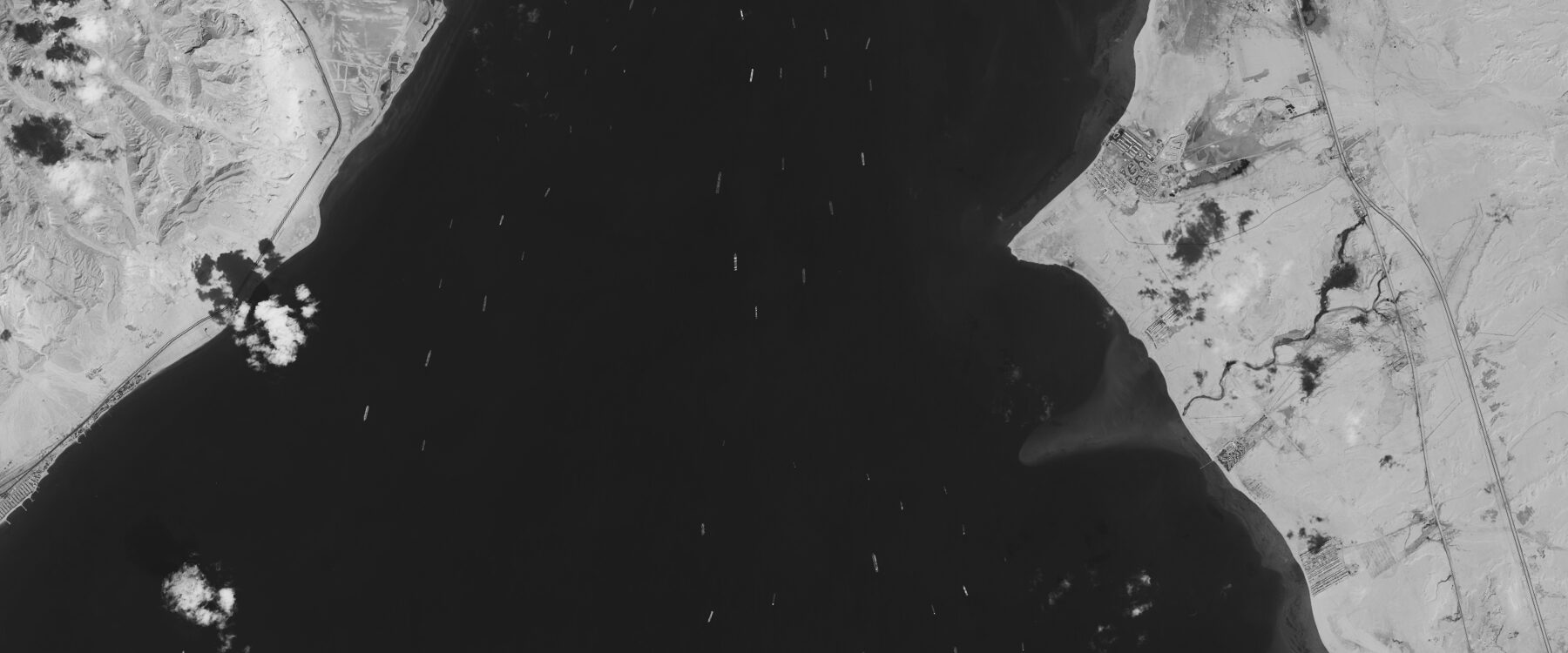 A grayscale satellite image of the Gulf of Suez: dozens of cargo ships and tankers lie at anchor as small pale marks across the dark water, with the desert coast, a road and a settlement along the eastern shore